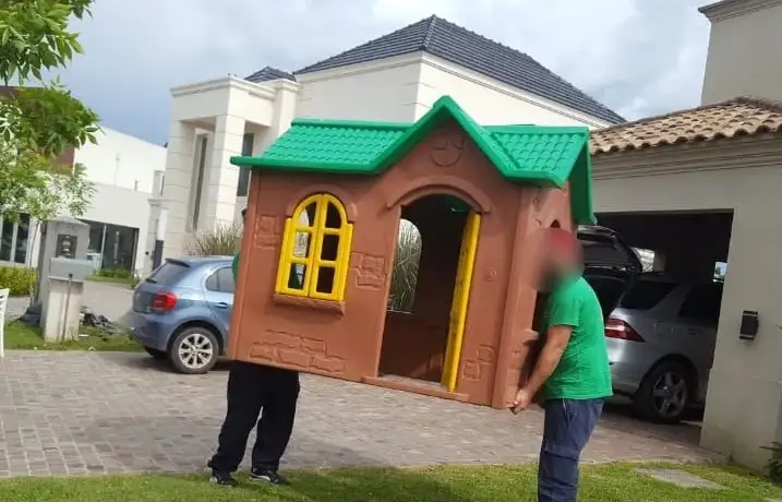 Traslado manual de una casita de juegos infantil de gran tamaño por el jardín durante una mudanza familiar.